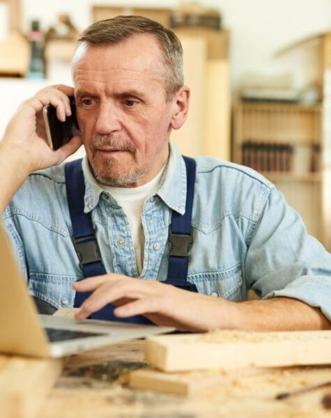 Portrait of modern senior carpenter speaking by phone and using laptop while working in joinery, copy space